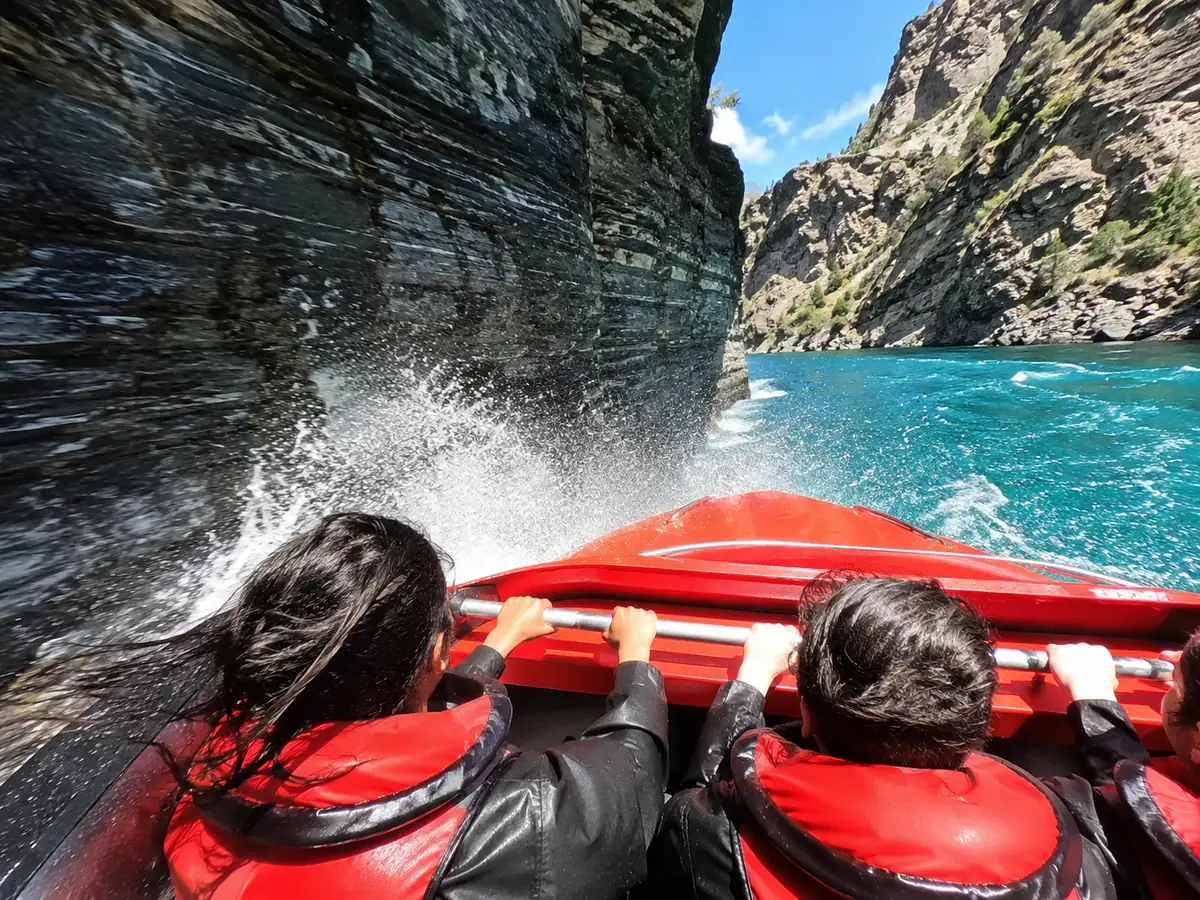 Riders gripping the rail as a red jet boat races past a sheer schist canyon wall