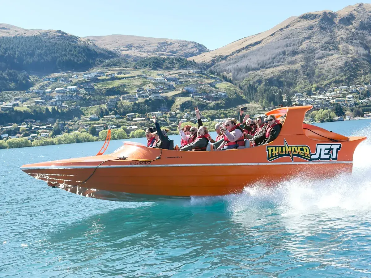 An orange jet boat speeding across Lake Wakatipu below snow-capped mountains